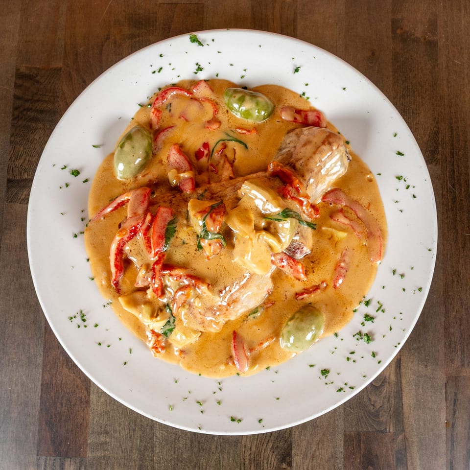 Creamy chicken dish with red peppers and green olives, garnished with parsley on a white plate.